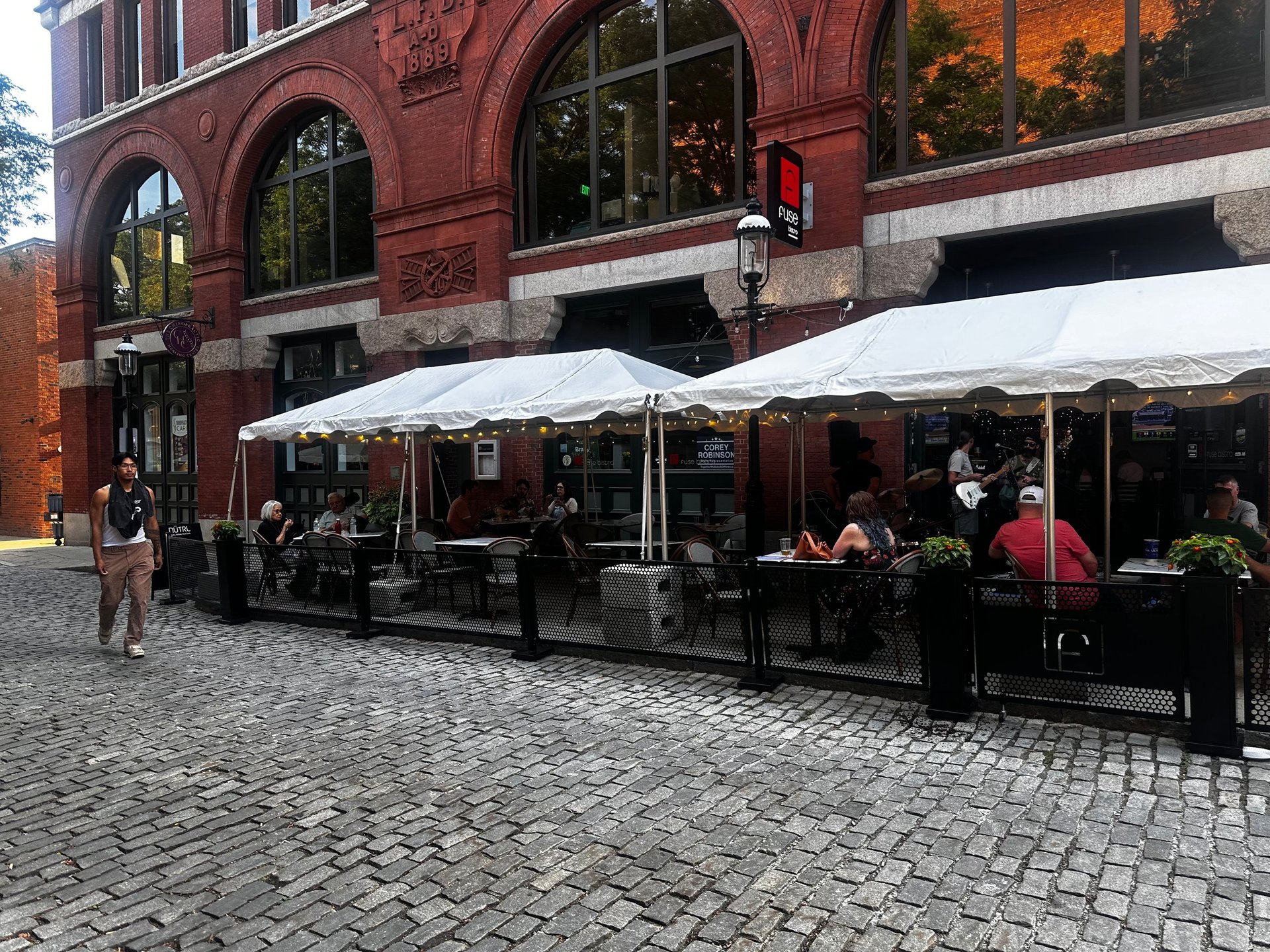 Outdoor cafe with white umbrellas along a cobblestone street in front of a historic red brick building