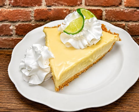 Slice of key lime pie with whipped cream and lime garnish on white plate against brick background
