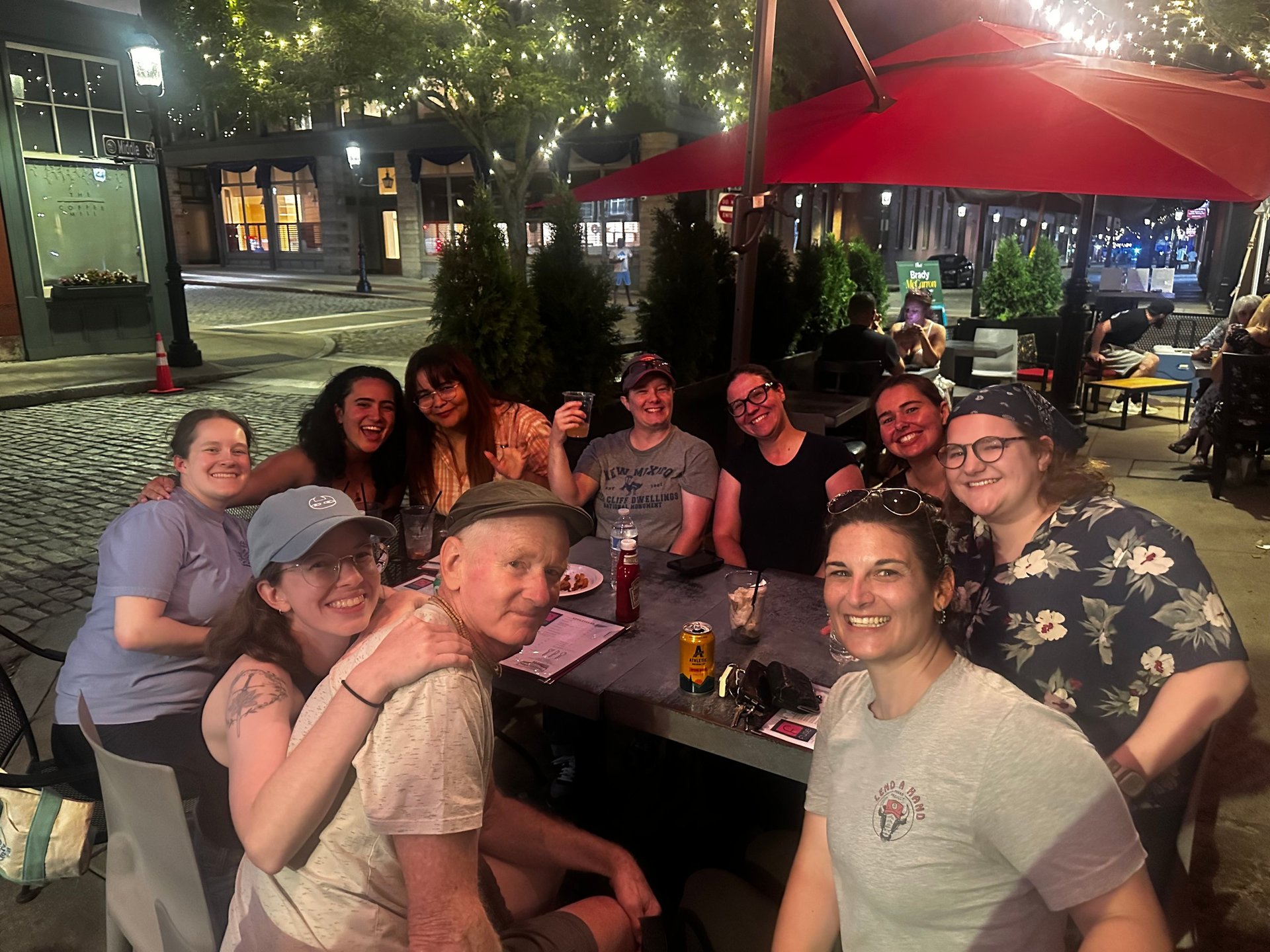 A group of friends gathered around a table at an outdoor night venue with red umbrellas and string lights overhead.